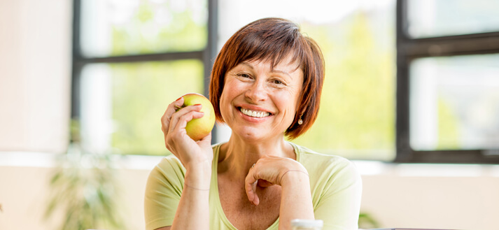 Dental Implants in College Station - woman with brown hair and brown eyes in a lime green shirt smiling while holding a yellow apple.