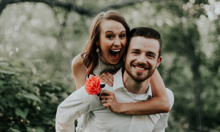 Brunette woman holds a coral flower in the forest while her brunette husband holds her on his back, both smiling