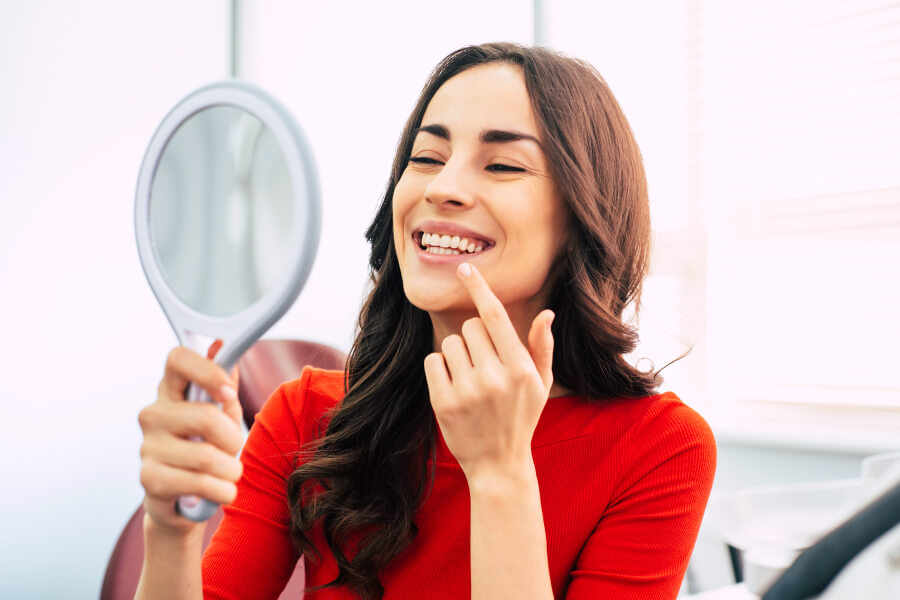 Brunette woman in a red blouse smiles in a mirror at her new smile after cosmetic dentistry